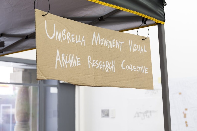 A piece of cardboard hung from a gazebo with the words “UMBRELLA MOVEMENT VISUAL ARCHIVE RESEARCH COLLECTIVE" written in white.