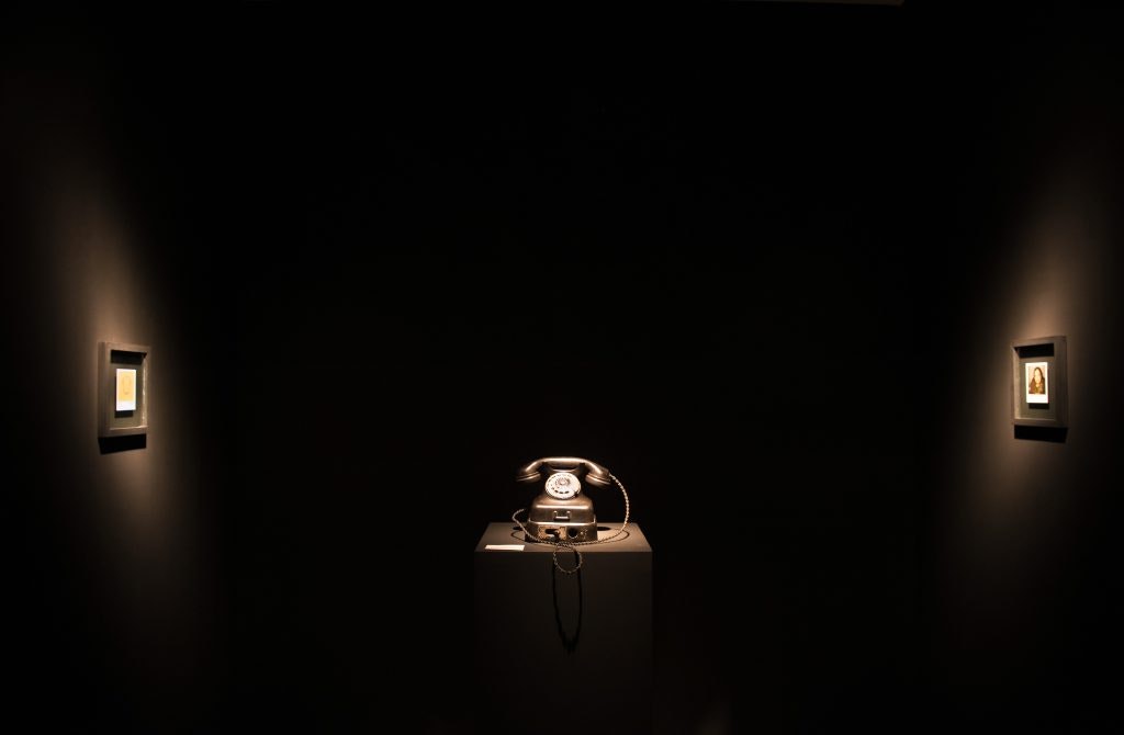 A telephone is lit up in a dark gallery, with two photoframes also lit up on the wall