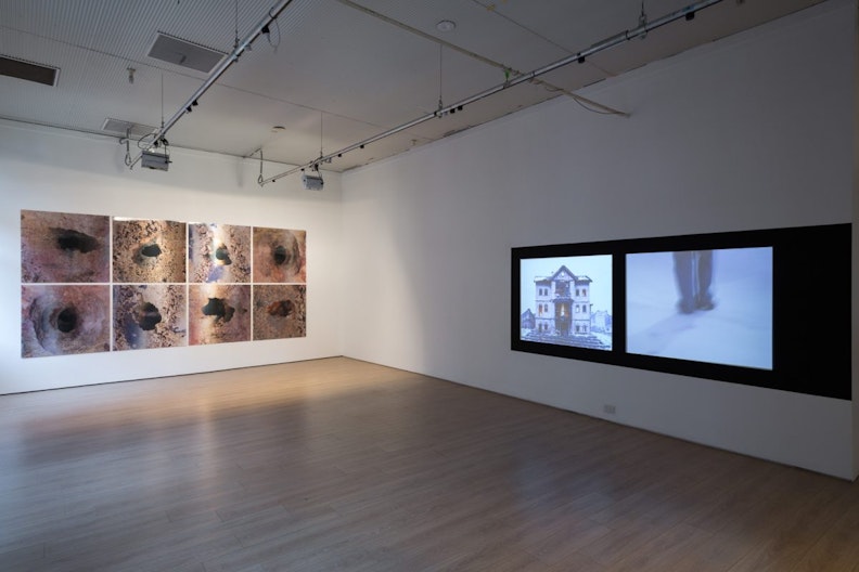 A gallery with eight etched copper artworks on the left wall, and two video screens showing a house and blurry legs next to each other
