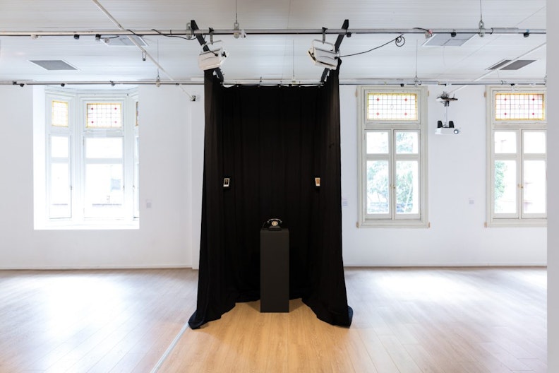 black curtains drape down from the centre of a brightly-lit gallery, inside is a black plinth with a telephone placed on top and two small picture frames on either side of the curtain