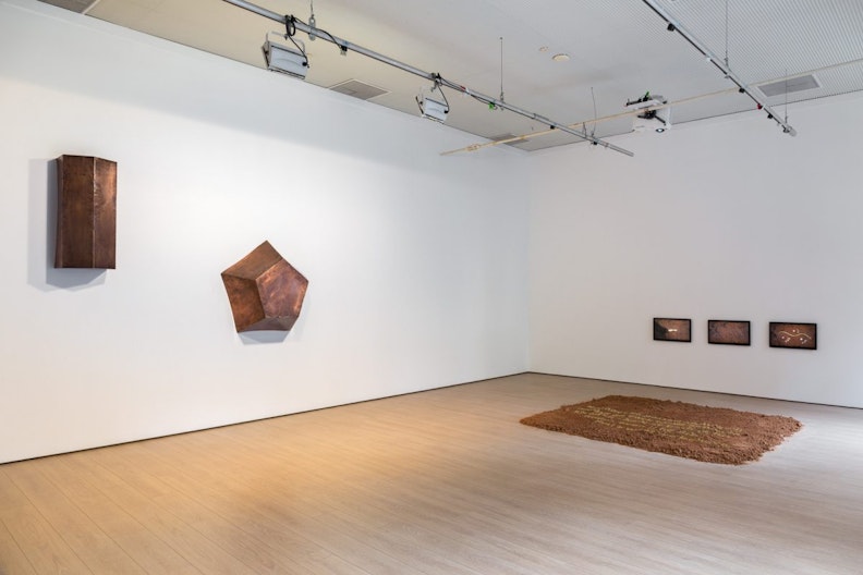 Two copper sculptures in the shape of a hexagonal cylinder and a pentagon are hung on different levels on the left side of the gallery wall, a patch of red earth rest on the ground to the right of the gallery, with three display screens sitting low on the wall