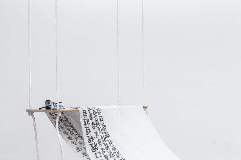 A fabric scroll is suspended on a wooden board with cotton strings. There are repeated Chinese letters written in ink on the scroll.