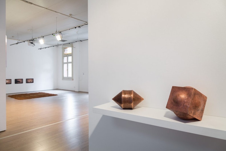 Two small copper sculptures in the shape of a double-edged cone and a cube with convex faces rest on a shelf at the entrance of the gallery