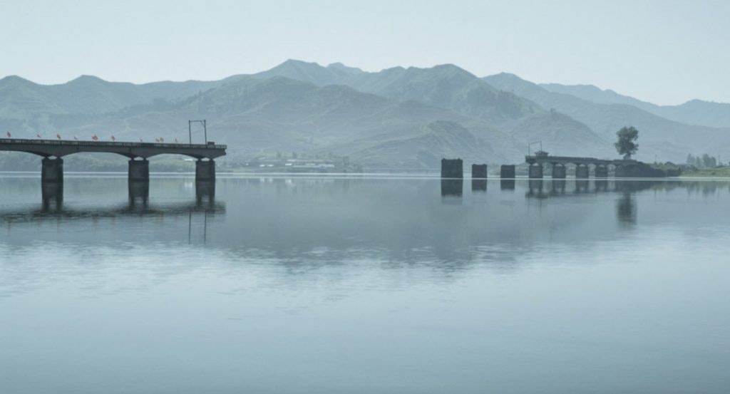 A film still from 'The Swim' showing a broken bridge over a lake reflecting misty mountains on the horizon