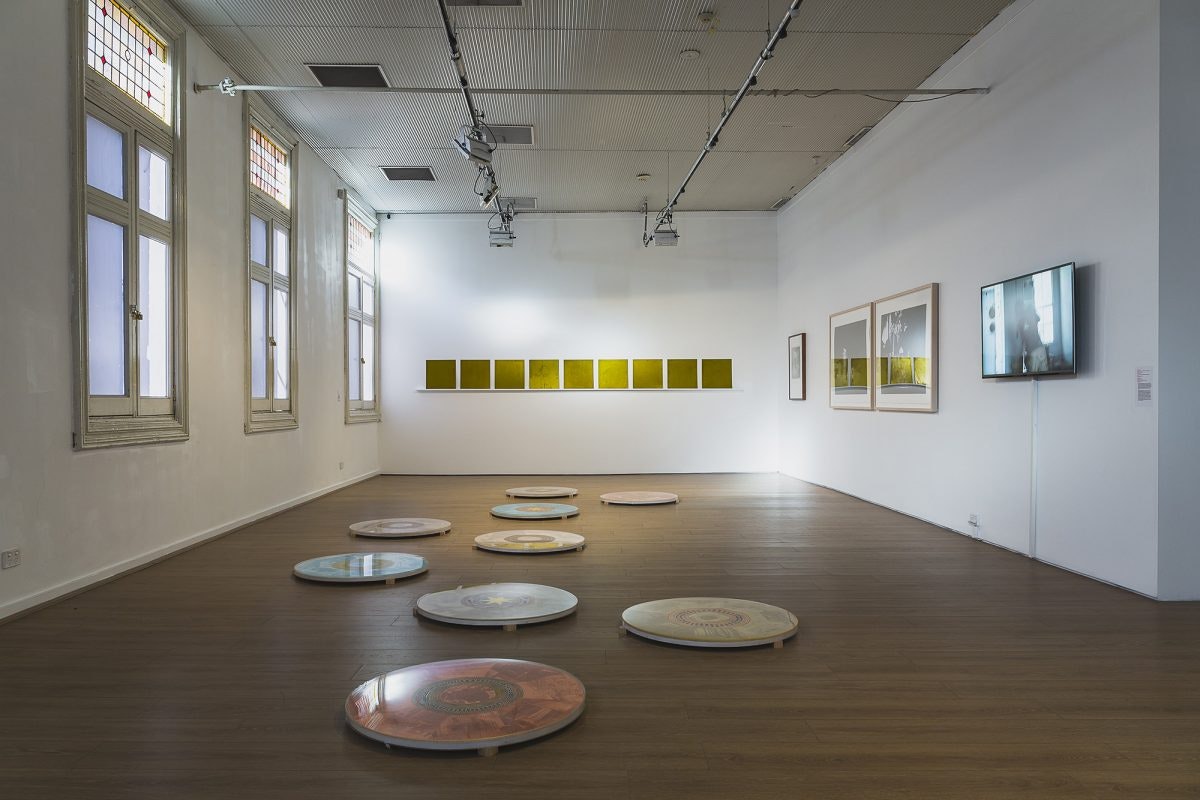 A gallery with a row of gold artworks on its far wall, framed photography prints and video artworks on its right wall, and shiny circle-shaped artworks on the ground