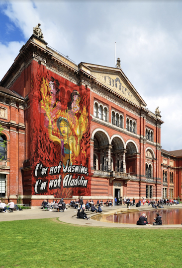 A daytime photo of the Victoria & Albert Museum, a decorative old London building. On half its main building’s entrance is a mural of a slender person with moustache, beautiful joined eyebrows and body hair, their skin in light-medium golden-amber tones. They wear a hairstyle and outfit deconstructed from Disney’s Jasmine and Aladdin - blue bandeau top, purple half-vest, blue headband with jewel, gold necklace and one chunky triangle earring, amber waistband and blue bottoms. They are engulfed from below waist down in red flames that also fill the background where red-toned Aladdin and Jasmine scream at the fire rising from the main figures' raised hands. In large block-cursive letters is the black/white title ‘I’m not Jasmine, I’m not Aladdin’