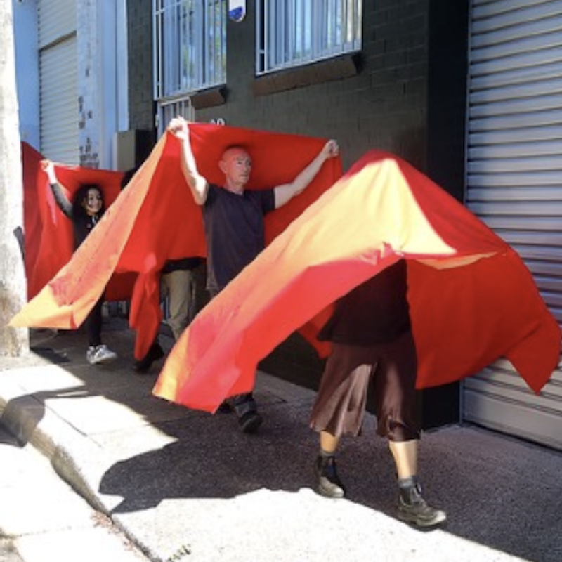 Four walkers with red fabric in a line walking along footpath; 1st walker has upper body covered in red cloth, second walker holds red cloth high above head which covers the third walker behind, and last walker is a little girl grinning, and holding the red cloth as high as possible above her head.