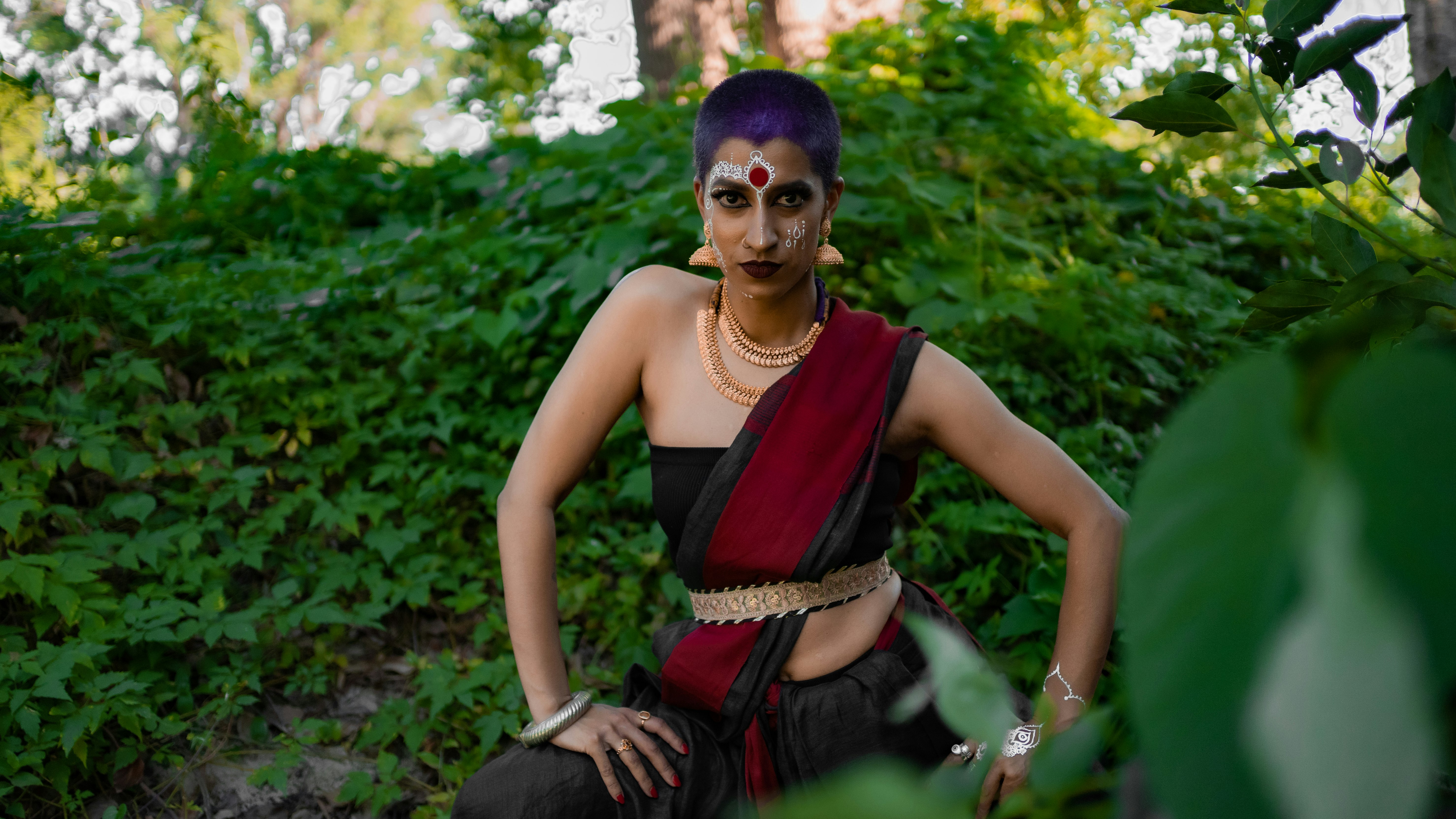 Shyamla is standing mid-movement with their hands on their thighs. They are wearing a strapless top and material draped like a sari over their shulder. They are adorned by gold jewellery, a pottu on their forehead and white mehndi on their face.