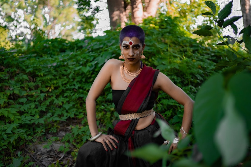 Shyamla is standing mid-movement with their hands on their thighs. They are wearing a strapless top and material draped like a sari over their shulder. They are adorned by gold jewellery, a pottu on their forehead and white mehndi on their face.