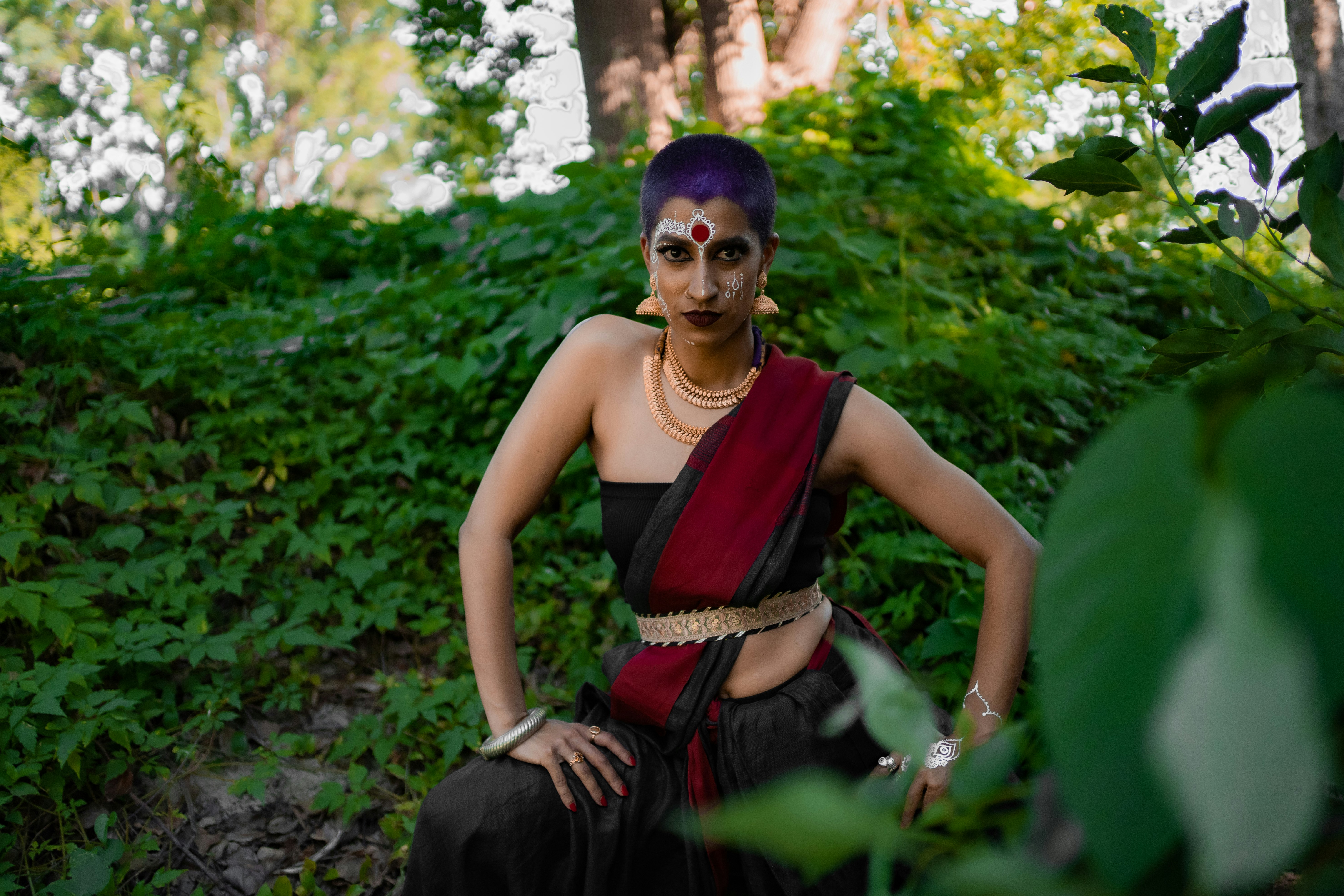 Shyamla is standing mid-movement with their hands on their thighs. They are wearing a strapless top and material draped like a sari over their shulder. They are adorned by gold jewellery, a pottu on their forehead and white mehndi on their face.