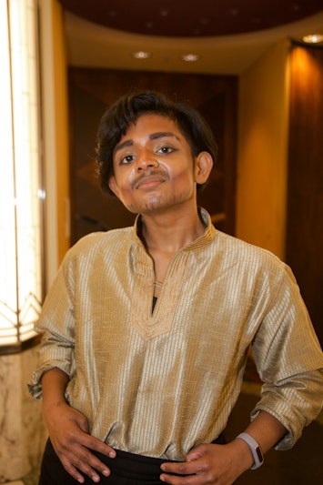 Manish is a Sri Lankan drag king with brown skin and short black hair. In this image he is looking into the camera and smiling slightly. He wears a gold kurta top tucked into black pants.