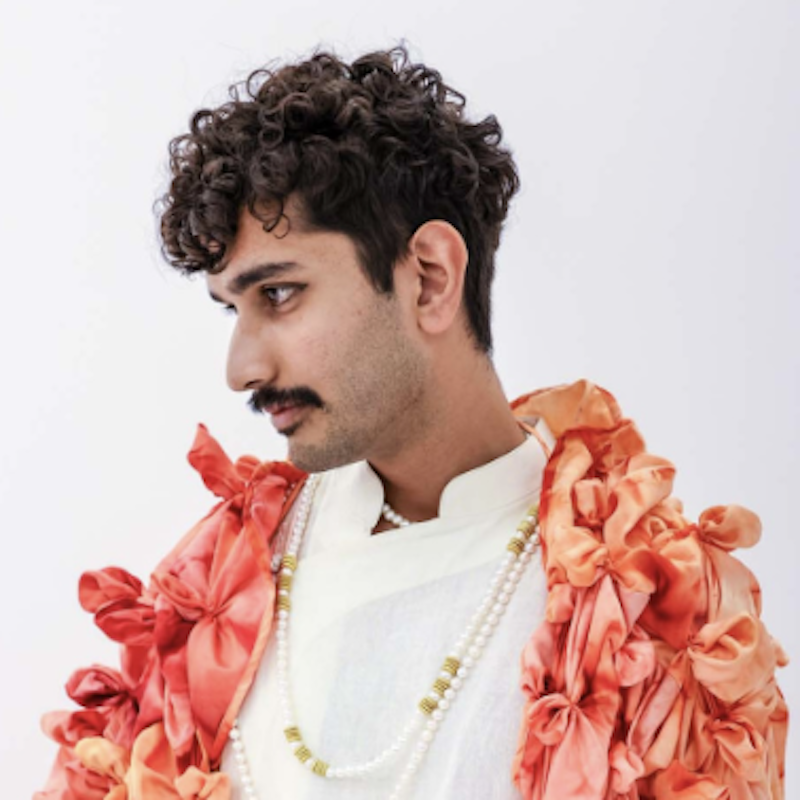 Rakish stands against a white background, looking off camera wearing a white Ladakhi Kurta and red coat with its material scrunched up to look like little flower petals hanging off the coat.