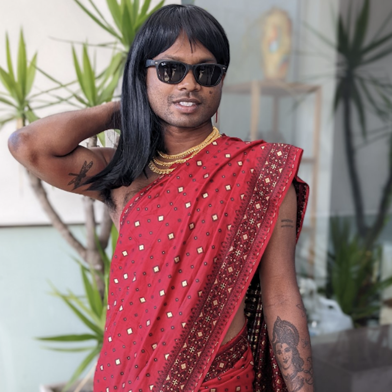 Salvin stands in front of the camera draped in a red sari with no blouse. Adorned with gold necklaces, wearing sunglasses and hand behind head, posing.