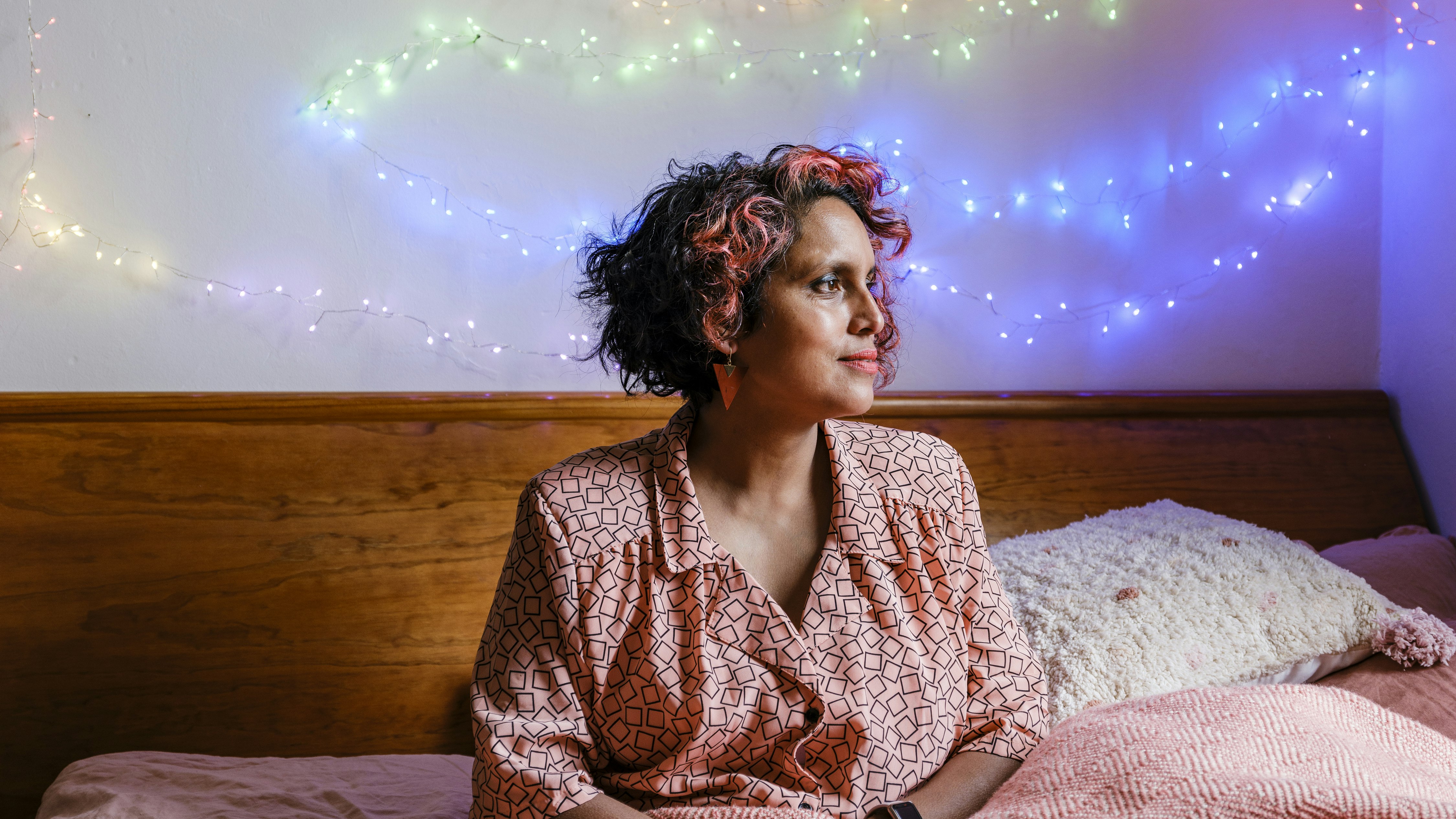 ID: daylight photo of a light-medium brown medium bodied person with coral, red and black hair, red drop earrings and lipstick and a coral shirt with thin black squares, sits in a peach blanketed bed with wooden headboard. Their face is in profile looking towards the light and delicate rainbow fairy lights are on the cream-coloured wall behind the bed.