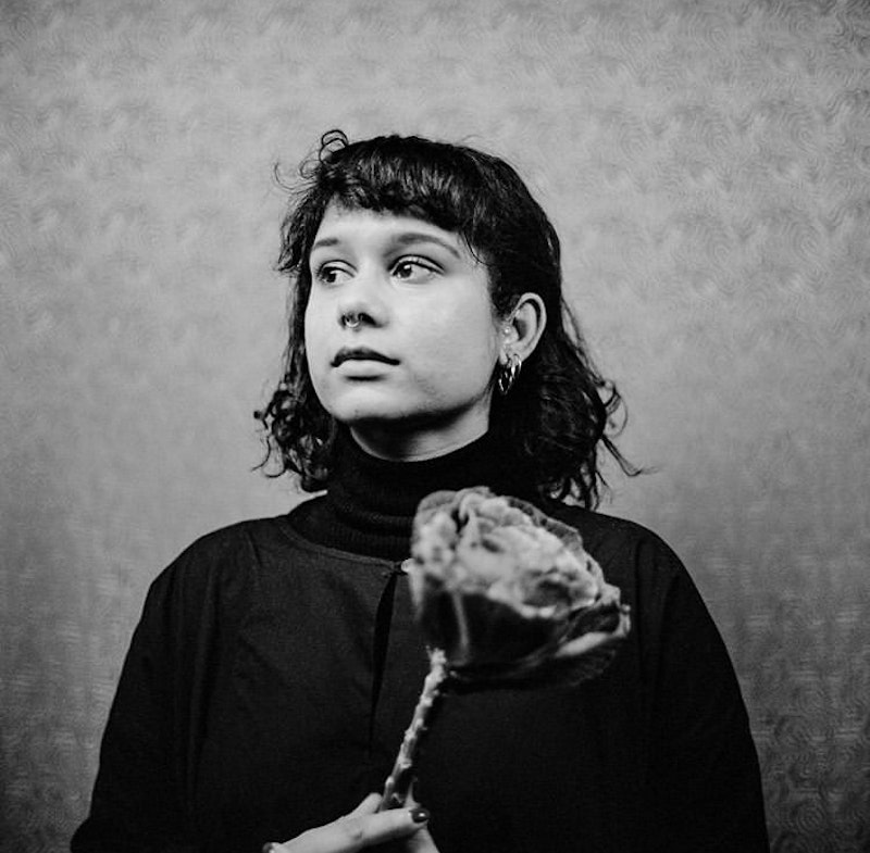 A black and white portrait of Shareeka. She is wearing a black dress layered over a black turtleneck. Her face is pensive and turned to the side, whilst clasping a cabbage flower. Her hair is black, mid length with a fringe and she is wearing some gold jewelry.