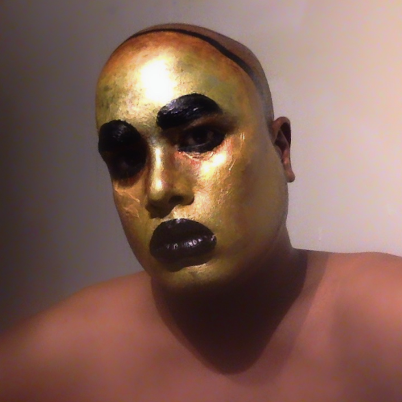 White background darkened on one side. Close-up of a brown body, shoulders to head. Bald Brown Face, gilded in dollar shop gold paint. Black lips. Dark eyes.