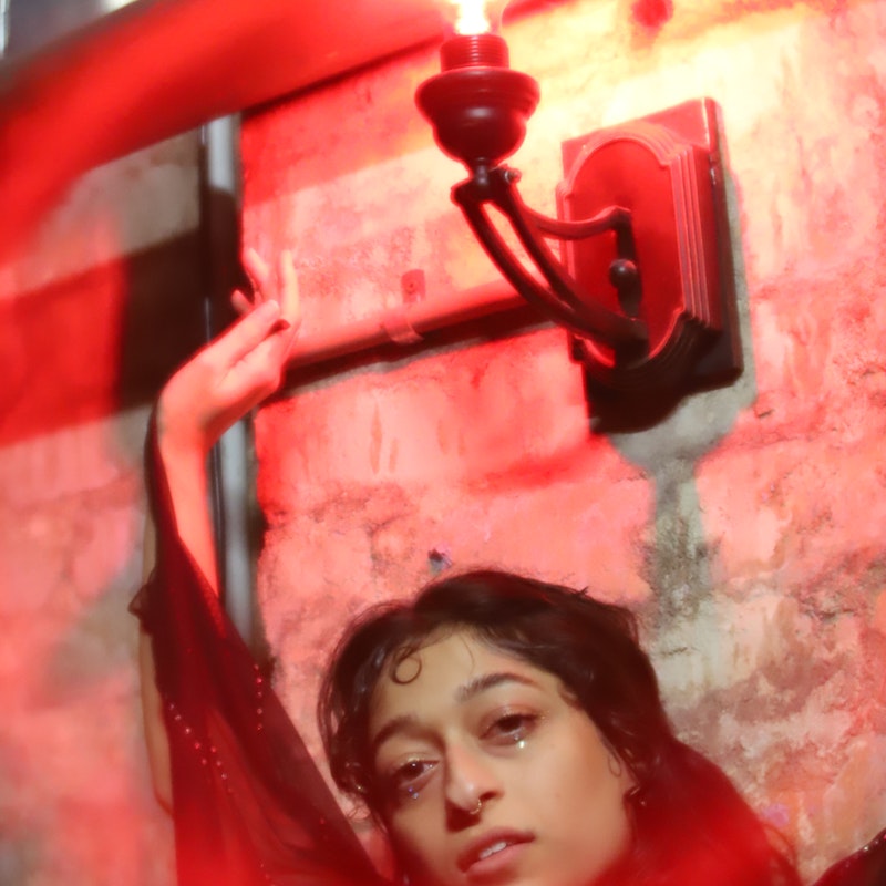 Salem is framed from the shoulders up in red lighting. they are wearing a bat-like sheer black cape that is rhinestoned, and they are holding one end of it up in the air. their have dark curly hair, and their expression is neutral and they have rhinestones glued underneath their eyes to match their outfit.