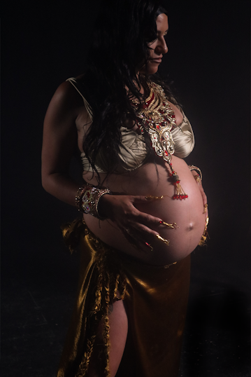 Paphilia stands in a dark space, holding her pregnant belly. She is wearing a gold bra, adorned with bold, illuminated jewellery. Her nails a long, embellished red and gold.