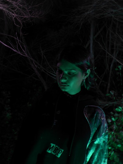 Image of a brown-skinned girl wearing a hoodie looking down at the ground, with a dark background and the leafless branches of a tree, and a green glow upon her.