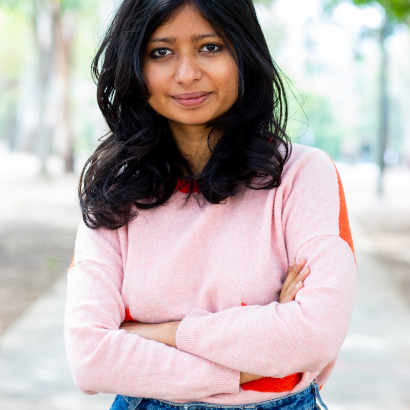 Brown-skinned woman with shoulder-length black hair, smiling at the camera, with her arms crossed. She is wearing a baby pink sweater and blue jeans and is standing outside, with trees behind her in soft focus.