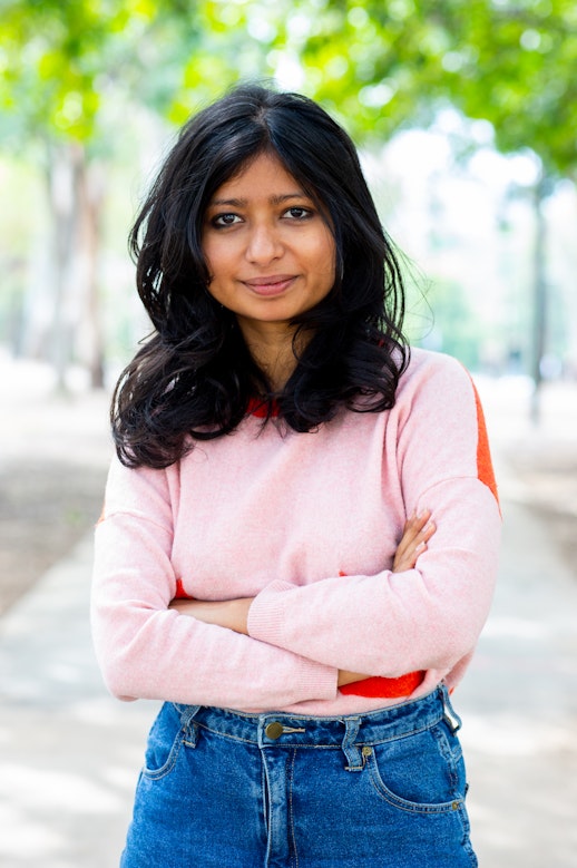 Brown-skinned woman with shoulder-length black hair, smiling at the camera, with her arms crossed. She is wearing a baby pink sweater and blue jeans and is standing outside, with trees behind her in soft focus.