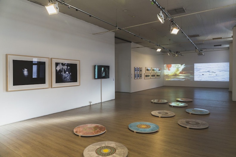 A gallery space with projected video on its far wall, framed photography prints on the left wall, and colourful circle artworks on the ground