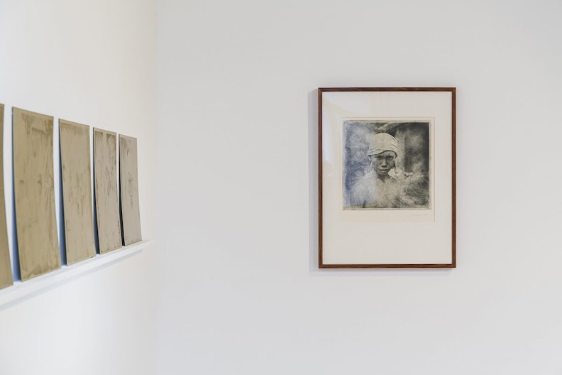 A corner of a gallery wall with a row of artworks sitting on a thin ledge on the left, and a front-on view of a square monochrome photograph in a retangle frame, showing a teenager with a serious expression