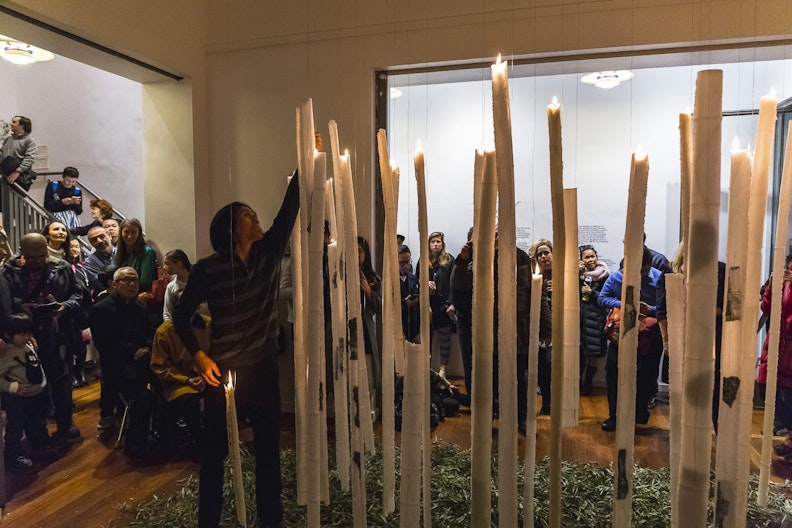 The artist, a man of medium height with dark hair, reaches up to light up the wax sculptures in the shape of segmented bamboo. Behind him, a crowd watches in the hallway and stairwell in the gallery