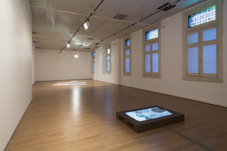 A partially-lit gallery with a video work on the floor and paper artworks at the far end of the gallery