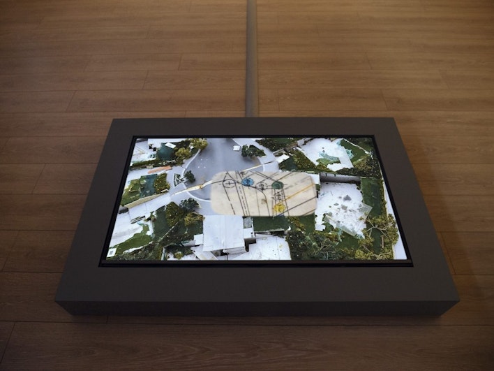 A video screen shows an aerial view of buildings amidst greenery, embedded in a wooden box