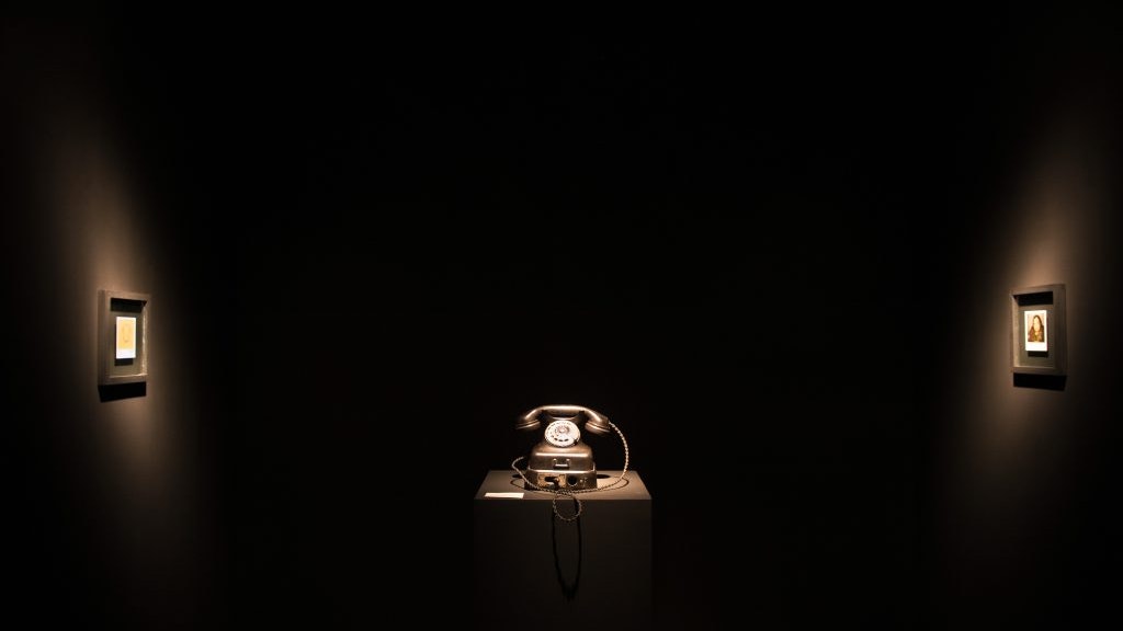 A dark room with a telephone on a stand in the centre and two framed images on dimly lit walls
