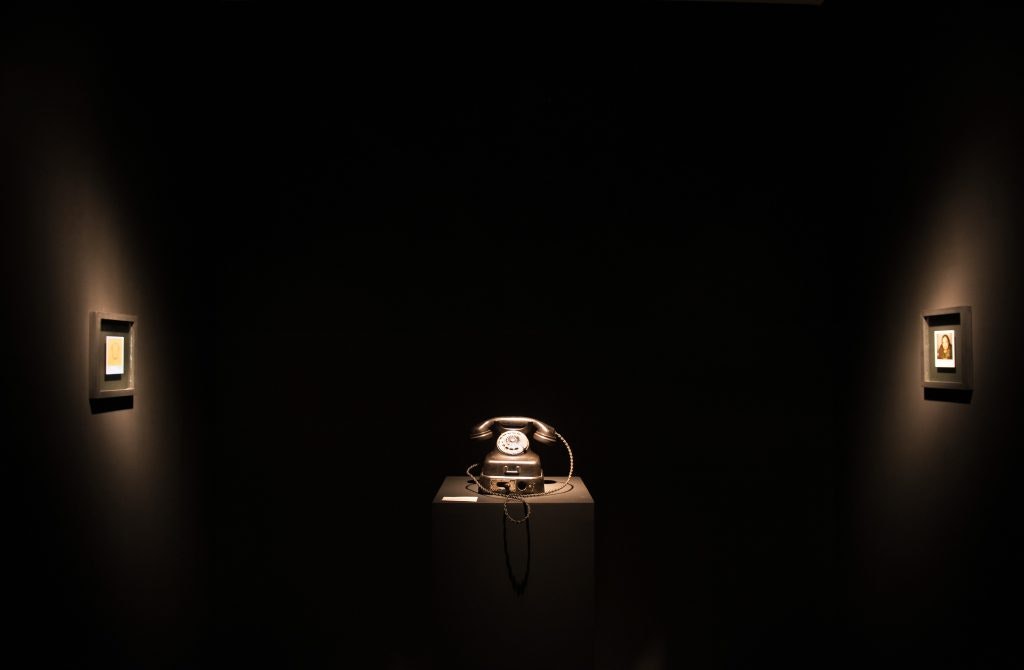 A dark room with a telephone on a stand in the centre and two framed images on dimly lit walls