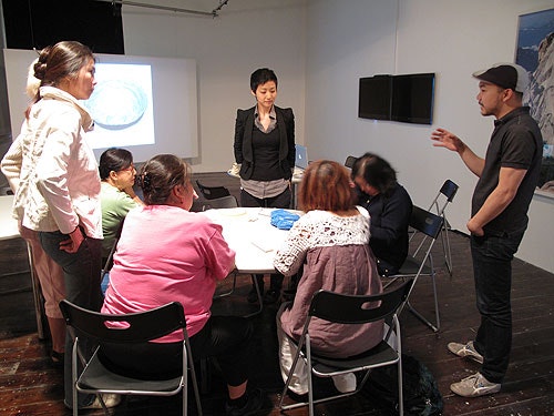 A group sits at a table during Liyen Chong's Threaded Stories Workshop and listens to a speaker
