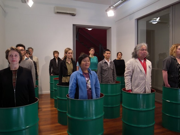 A photo showing Tatsumi Orimoto's Oil Can performance with people standing motionless in green oil cans, at 4A Centre