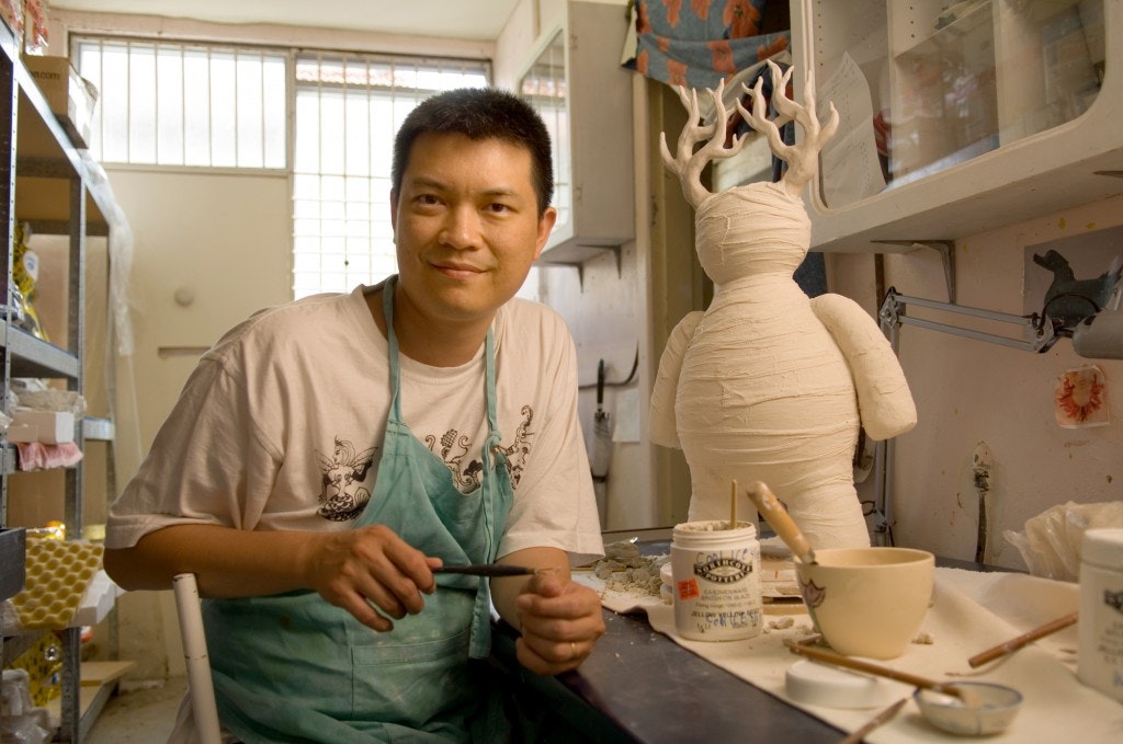 A photo of Vipoo Srivilasa sitting at a workshop table with a sculpture