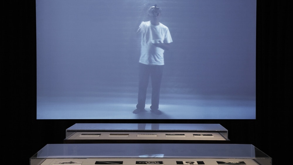 A screen showing a person standing alone, in front of the screen are two glass displays