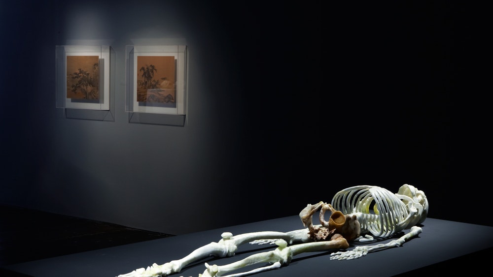 A photo of a human skeleton lying on a black platform as part of He Xiangyu's exhibition Cola Project