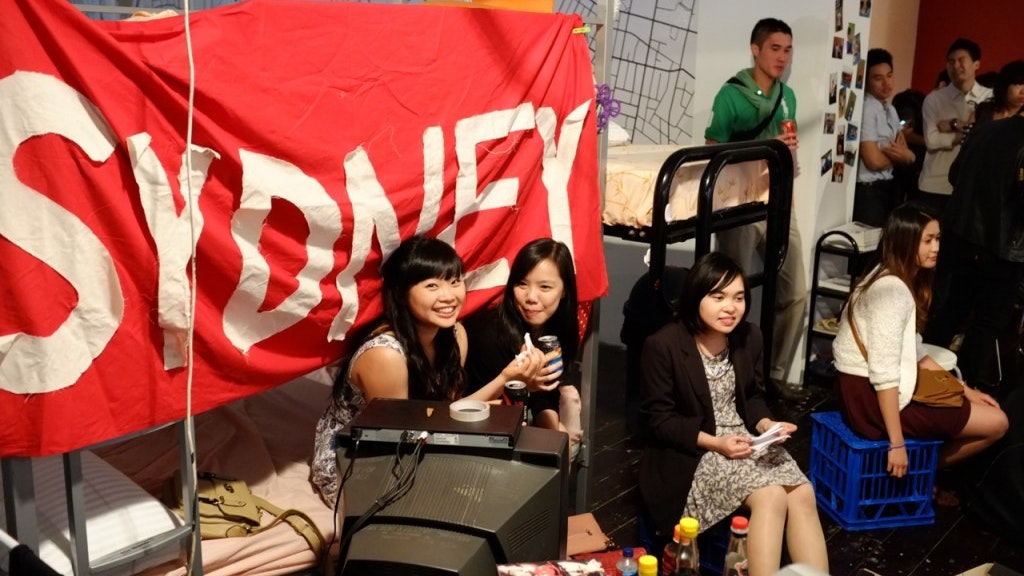 A photograph showing people sitting and standing at a party with a red banner that reads Sydney in white text