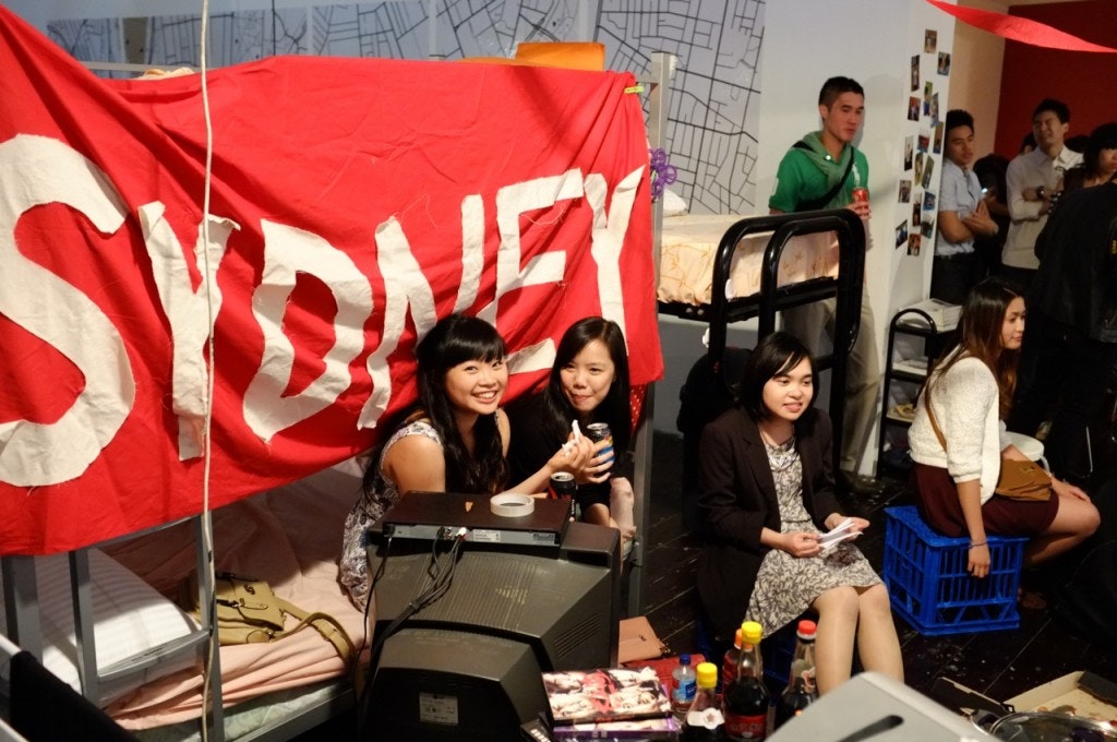 A photograph showing people sitting and standing at a party with a red banner that reads Sydney in white text