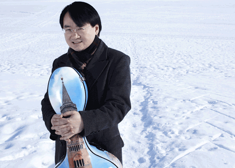 A photo of cellist Jian Wang standing in the snow holding a cello case