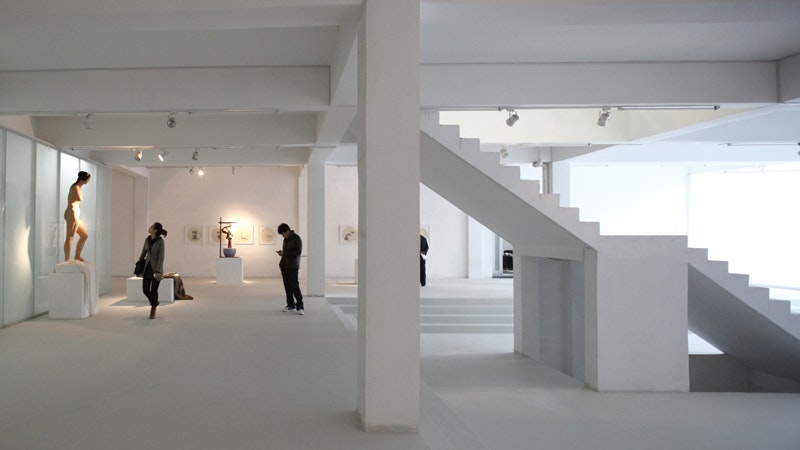 The white interior of an art gallery with a staircase and two people examining artworks