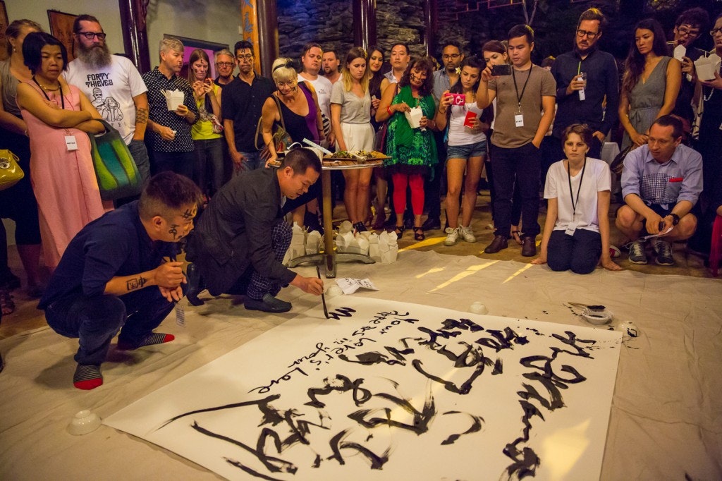 A photo of two artists creating shu fa (Chinese calligraphy) on a sheet with a large crowd gathered watching at night