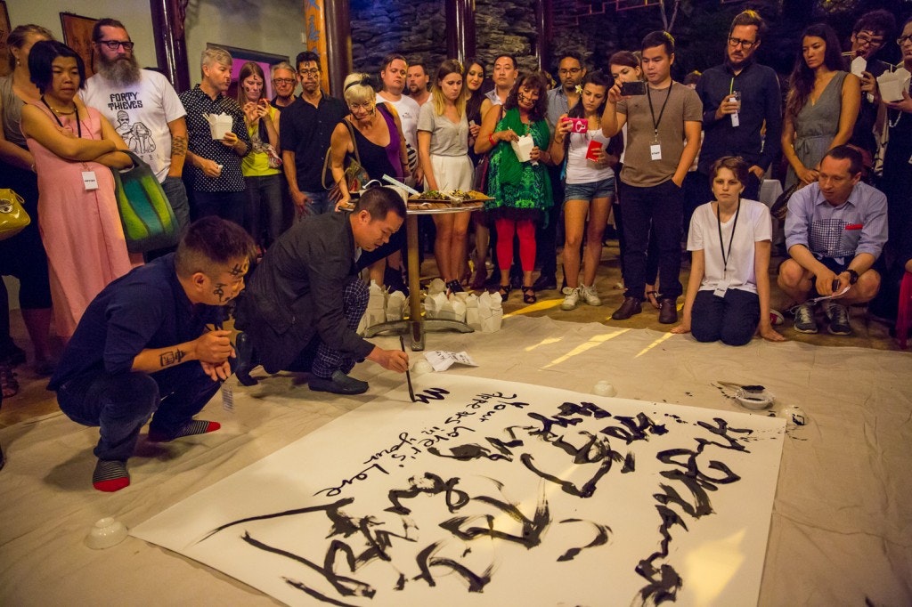 A photo of two artists creating shu fa (Chinese calligraphy) on a sheet with a large crowd gathered watching at night