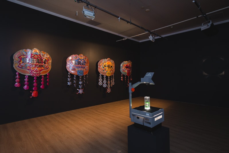 Wall: Abdullah M.I. Syed, Brut for Men series (2013), hand-beaten metal medallion and chamak patti (hand-made sticker ornamentation), wood, stainless steel; installation view, 4A Centre for Contemporary Asian Art.  Centre: Abdullah M.I. Syed, The Fragrance of the Moon (2013), Brut for Men bottle, Perspex and transparency projector, installation view, 4A Centre for Contemporary Asian Art. Courtesy the artist. Image: Document Photography.