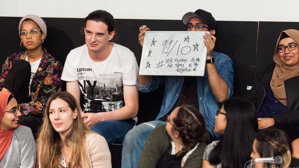 Audience members watching a poetry slam, with one person holding up a sign with scores.