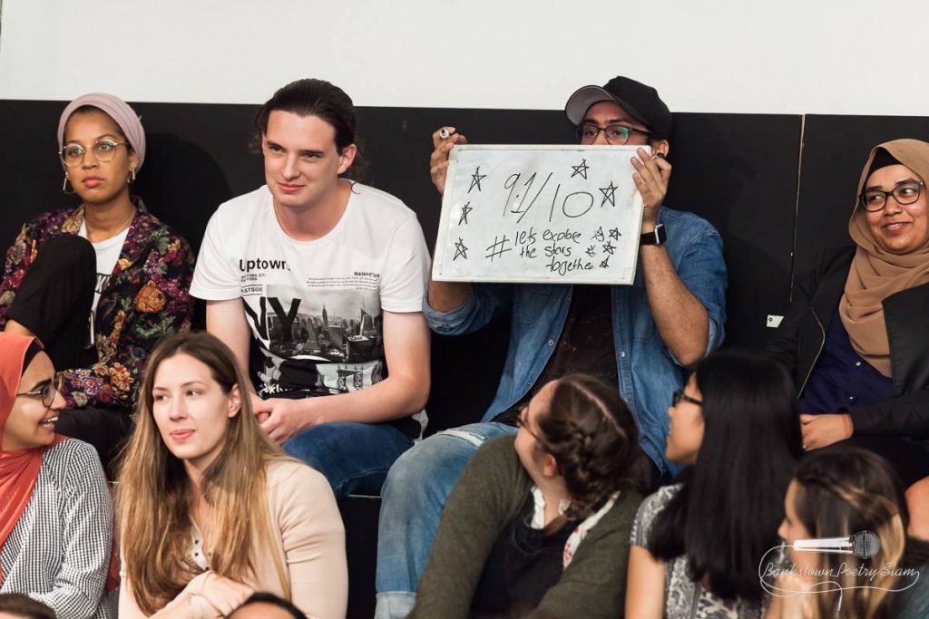 Audience members watching a poetry slam, with one person holding up a sign with scores.