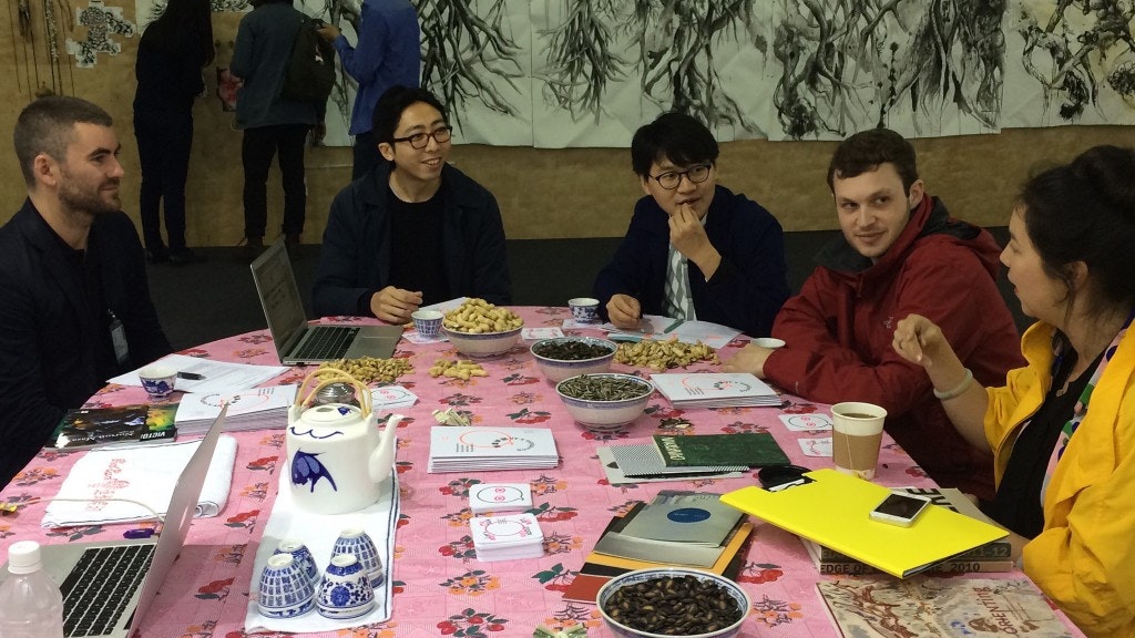 Five people sitting at a table covered by books and food with an artwork hanging against the far wall.