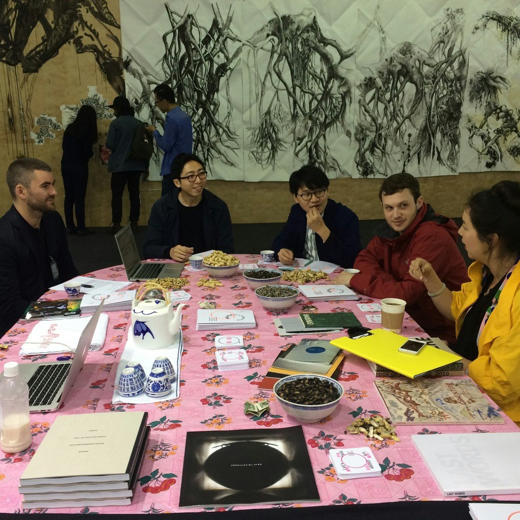 Five people sitting at a table covered by books and food with an artwork hanging against the far wall.