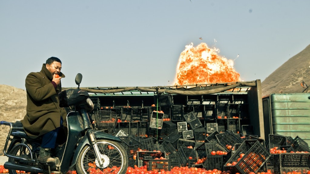 A film still depicting a man riding a motorcycle and biting a tomato, with scattered crates of tomatoes and an explosion in the background.