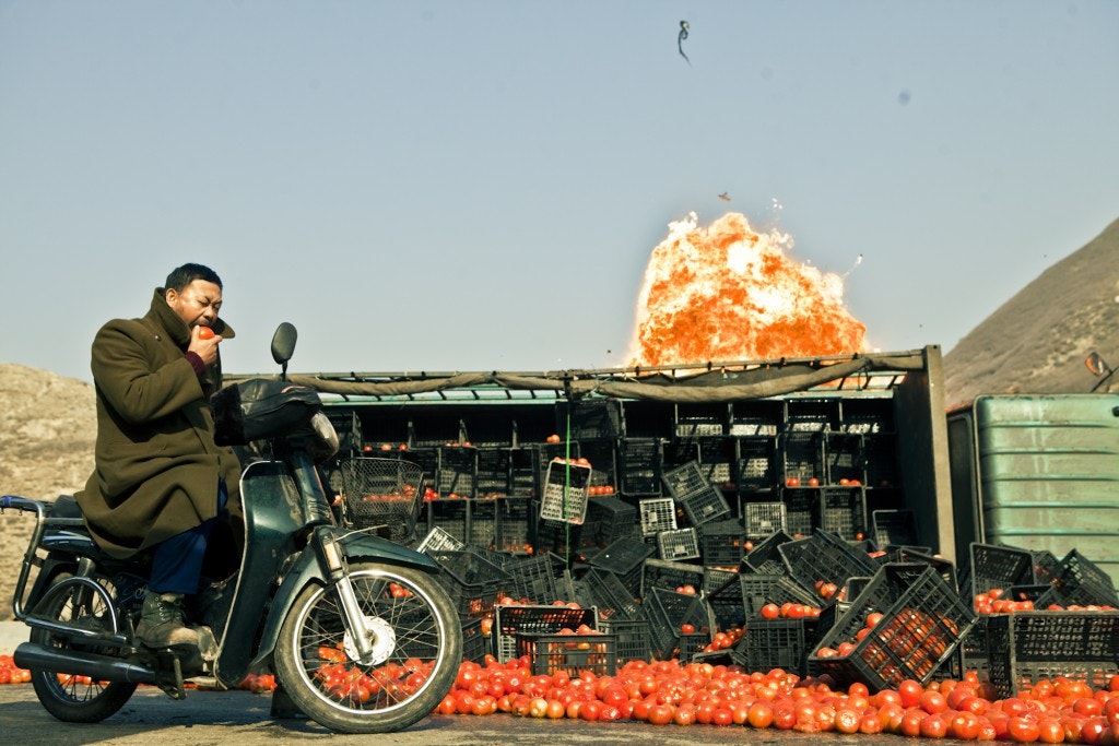 A film still depicting a man riding a motorcycle and biting a tomato, with scattered crates of tomatoes and an explosion in the background.